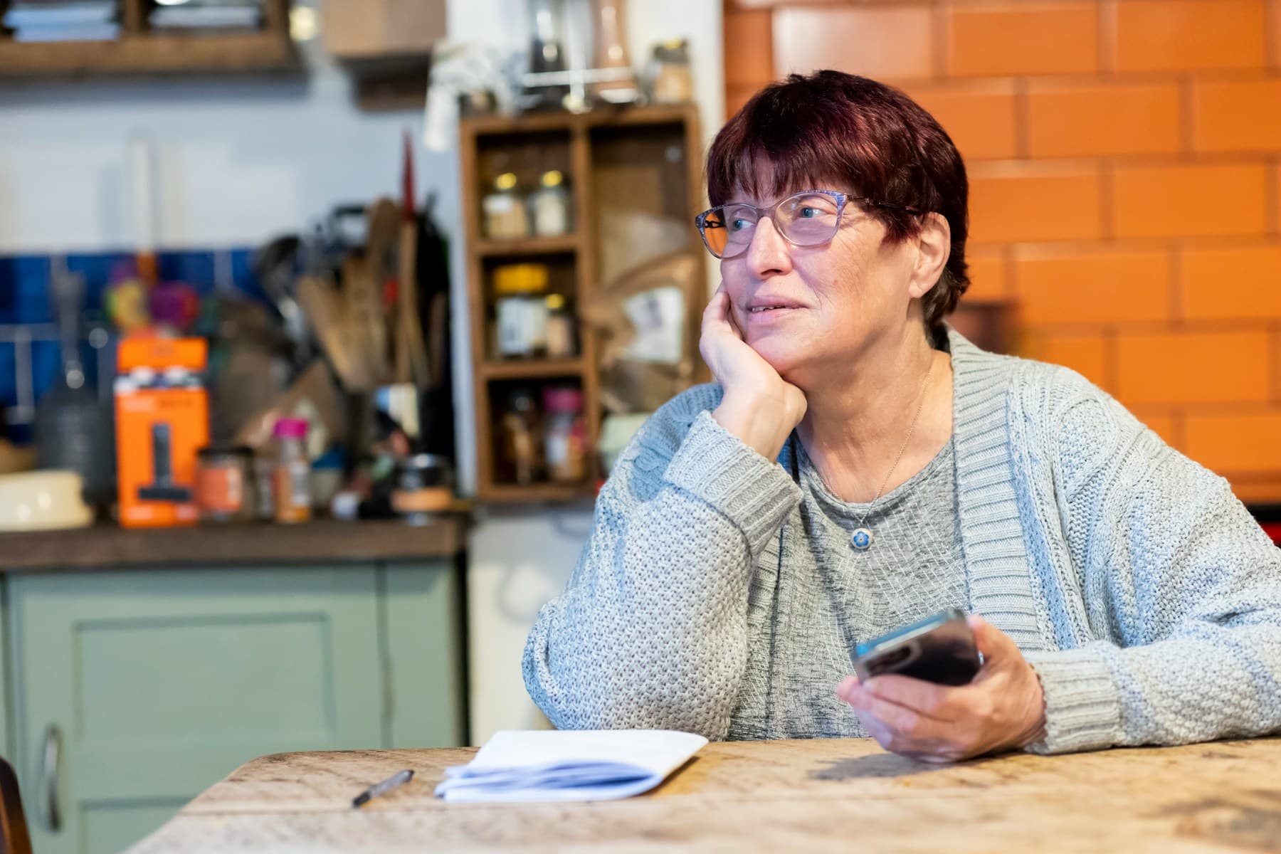 Une femme âgée, téléphone en main, réfléchit assise à la table de sa cuisine, un carnet ouvert devant elle.